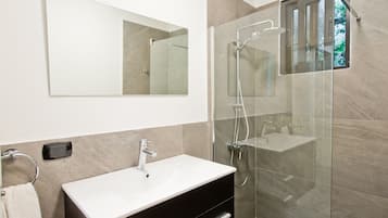 Un baño moderno con lavamanos blanco, mueble oscuro y ducha con cerramiento de vidrio.