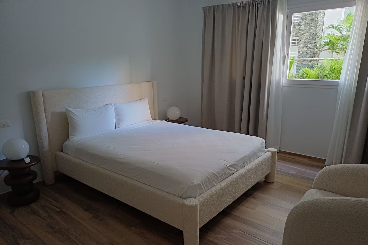 A neatly made bed with white linens in a room with wooden flooring, a nightstand with a lamp, and a view of greenery through a window with curtains.