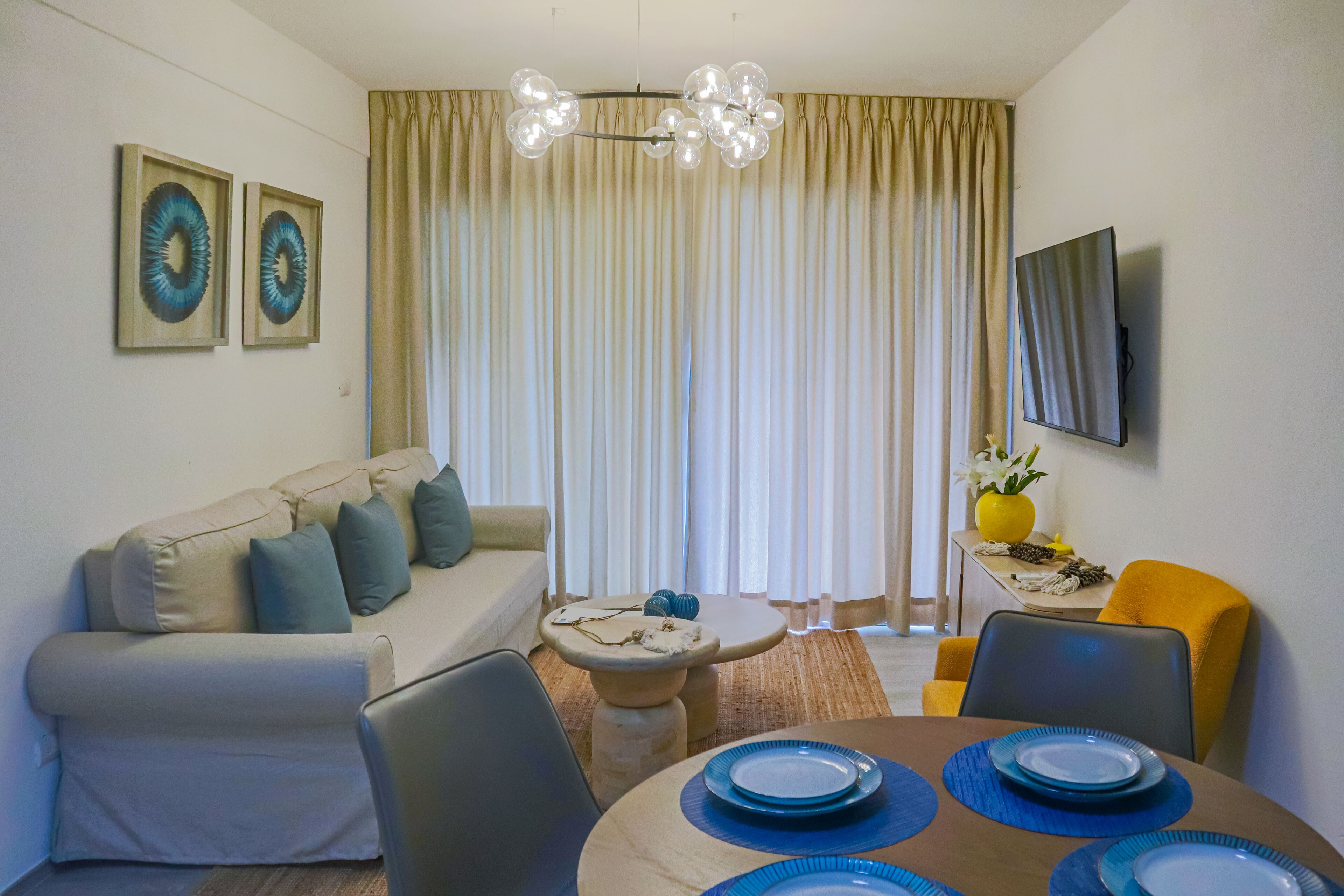 Una sala moderna con un sofá, una mesa de comedor y sillas. Hay un televisor montado en la pared y una ventana con cortinas.