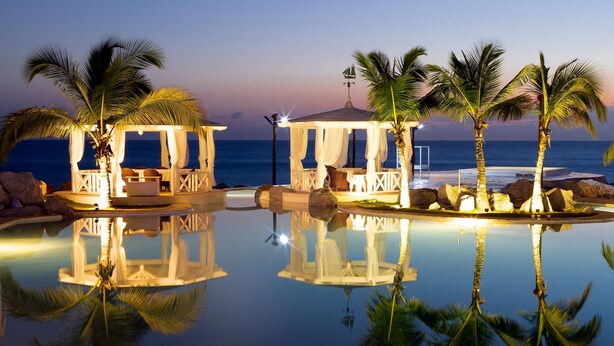 A serene pool area with two covered seating areas, palm trees, and a clear view of the ocean at dusk. - TRACADERO Beach Club and Holidays Rentals (San Rafael del Yuma)