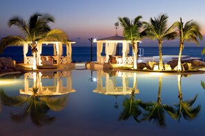 A serene pool area with two covered seating areas, palm trees, and a clear view of the ocean at dusk.