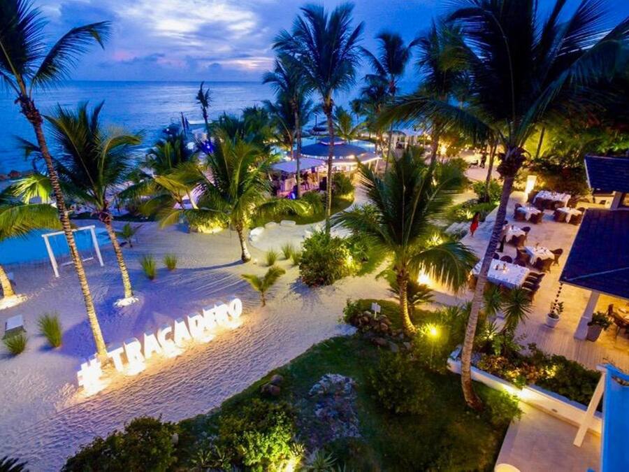 A resort with a beach area, palm trees, and a sign reading "TRAGACOMA".