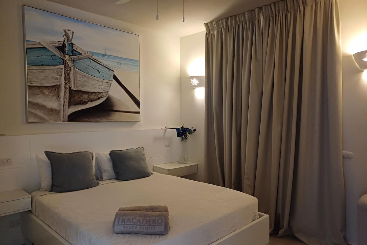 A neatly made bed with a towel labeled "TRACADERO BEACH RESORT" and a painting of a boat on the wall.