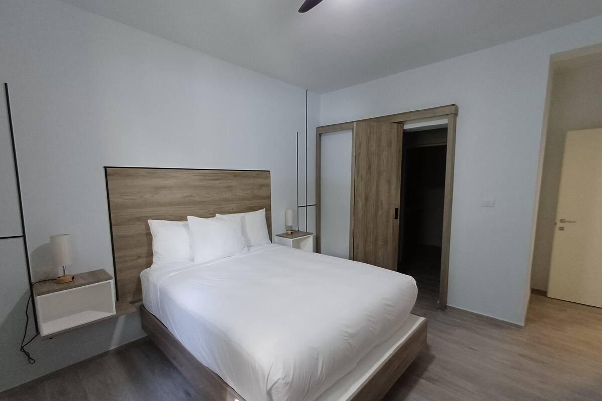 A modern bedroom with a wooden headboard, a bedside table with a lamp, and a wardrobe.