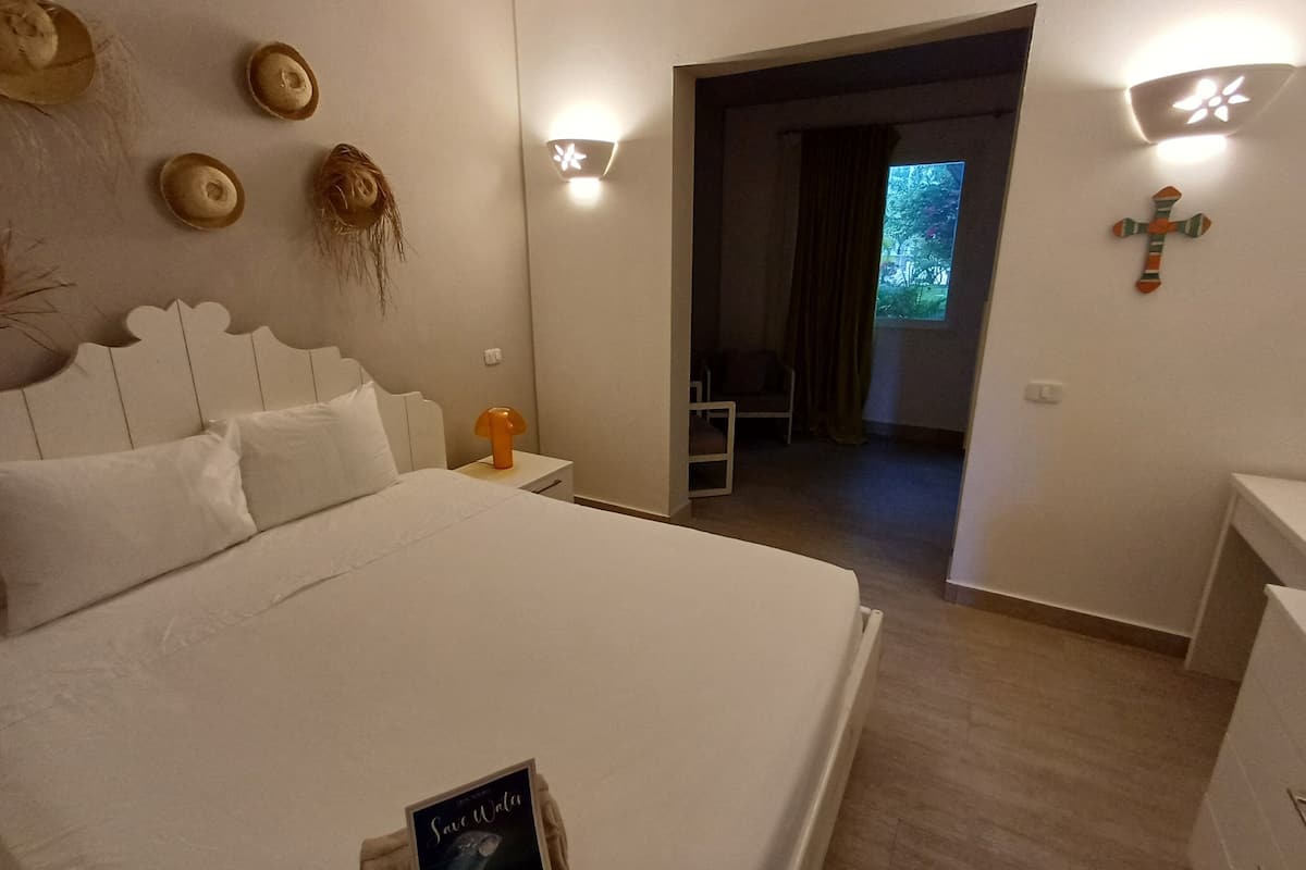 A bedroom with a large bed, a bedside table with a lamp, a small table with a book, and a cross on the wall.