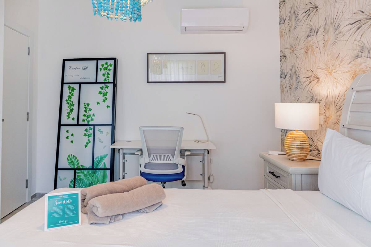 A hotel room with a bed, a desk with a chair, a nightstand with a lamp, and a wall-mounted air conditioner.