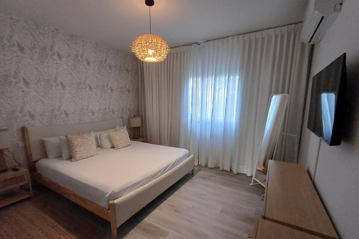 A bedroom with a bed, a bedside table, a lamp, a TV, and a window with curtains.