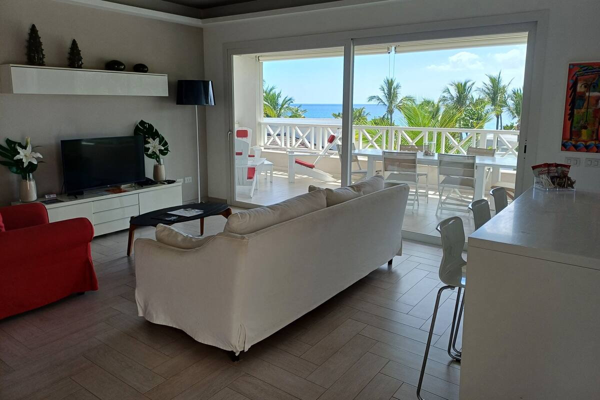A modern living room with a white sofa, red armchair, and a flat-screen TV on a white console. A balcony with white chairs and a table is visible, overlooking a tropical landscape.