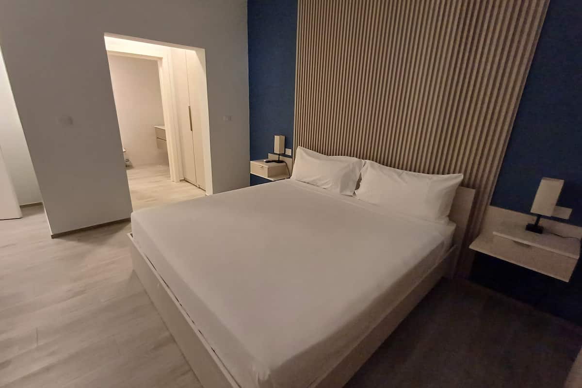 A modern hotel room with a large bed, a bedside table with a lamp, and a view of the hallway.