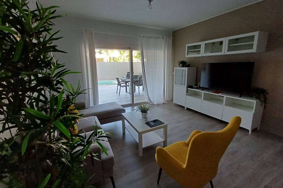 A living room with a sofa, a yellow armchair, a coffee table, and a TV on a white cabinet. There is a sliding glass door leading to an outdoor area with a table and chairs.