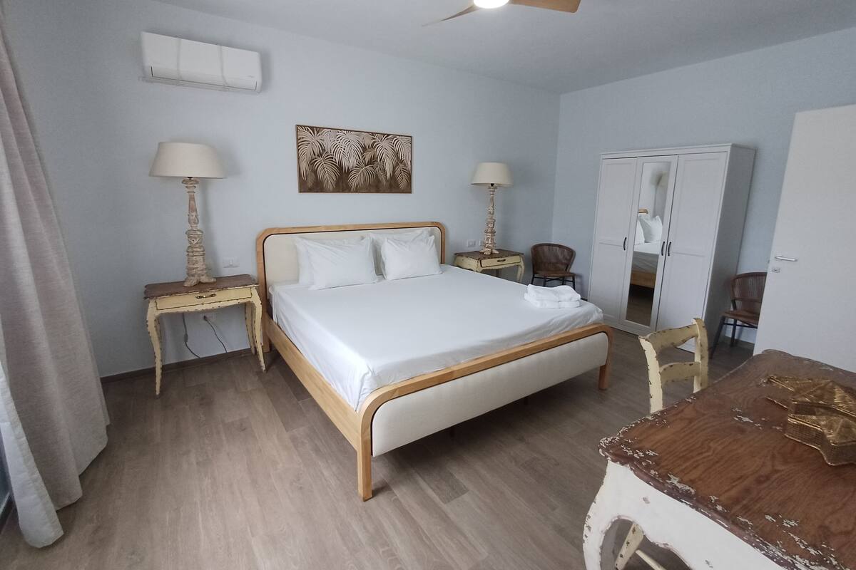 A hotel room with a bed, two bedside tables, a ceiling fan, a wardrobe, and a small table with a lamp.