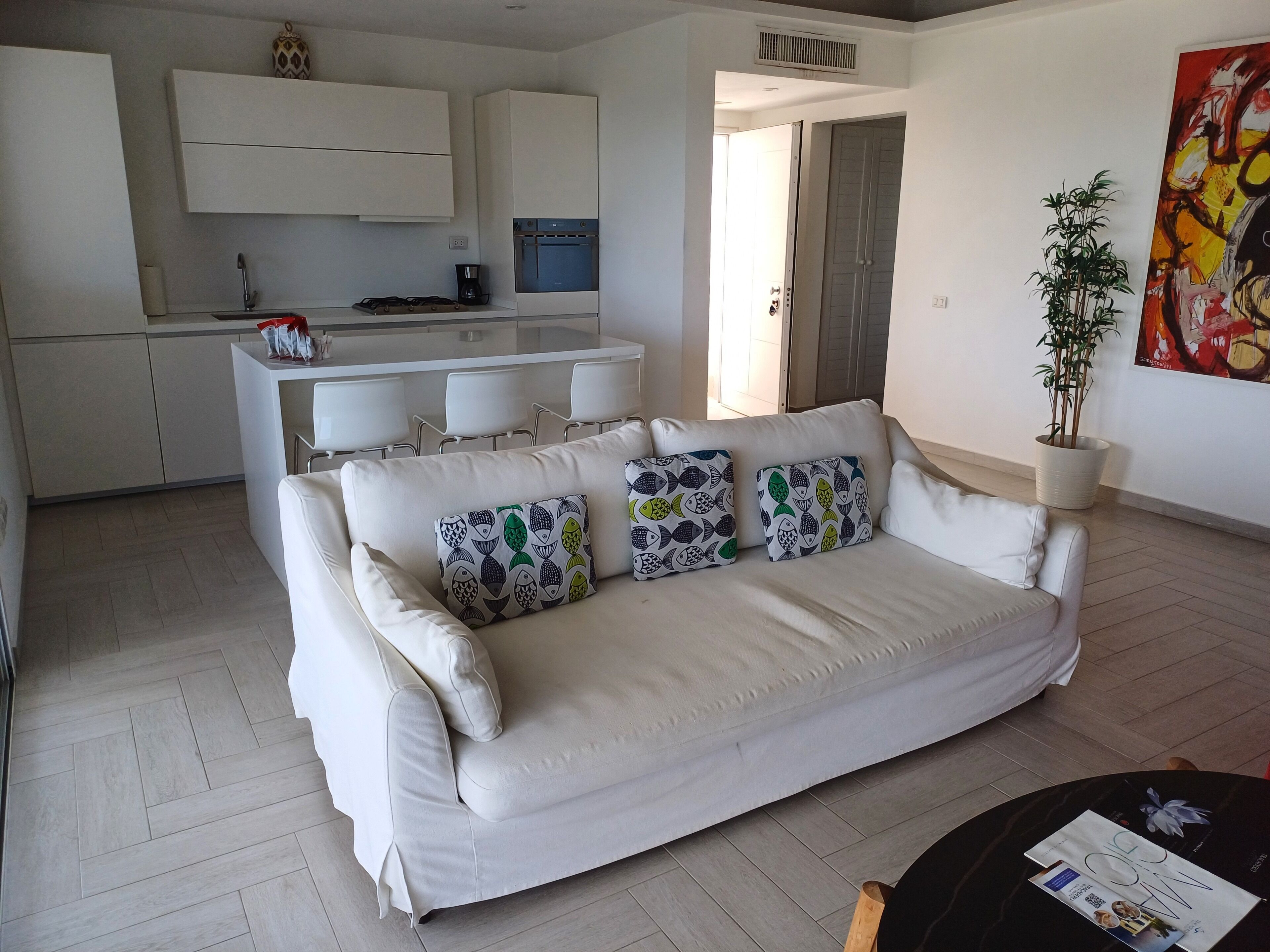 Ein modernes Wohnzimmer mit einem weißen Sofa, gemusterten Kissen, einer Kaffeemaschine und einem Essbereich mit weißen Stühlen.