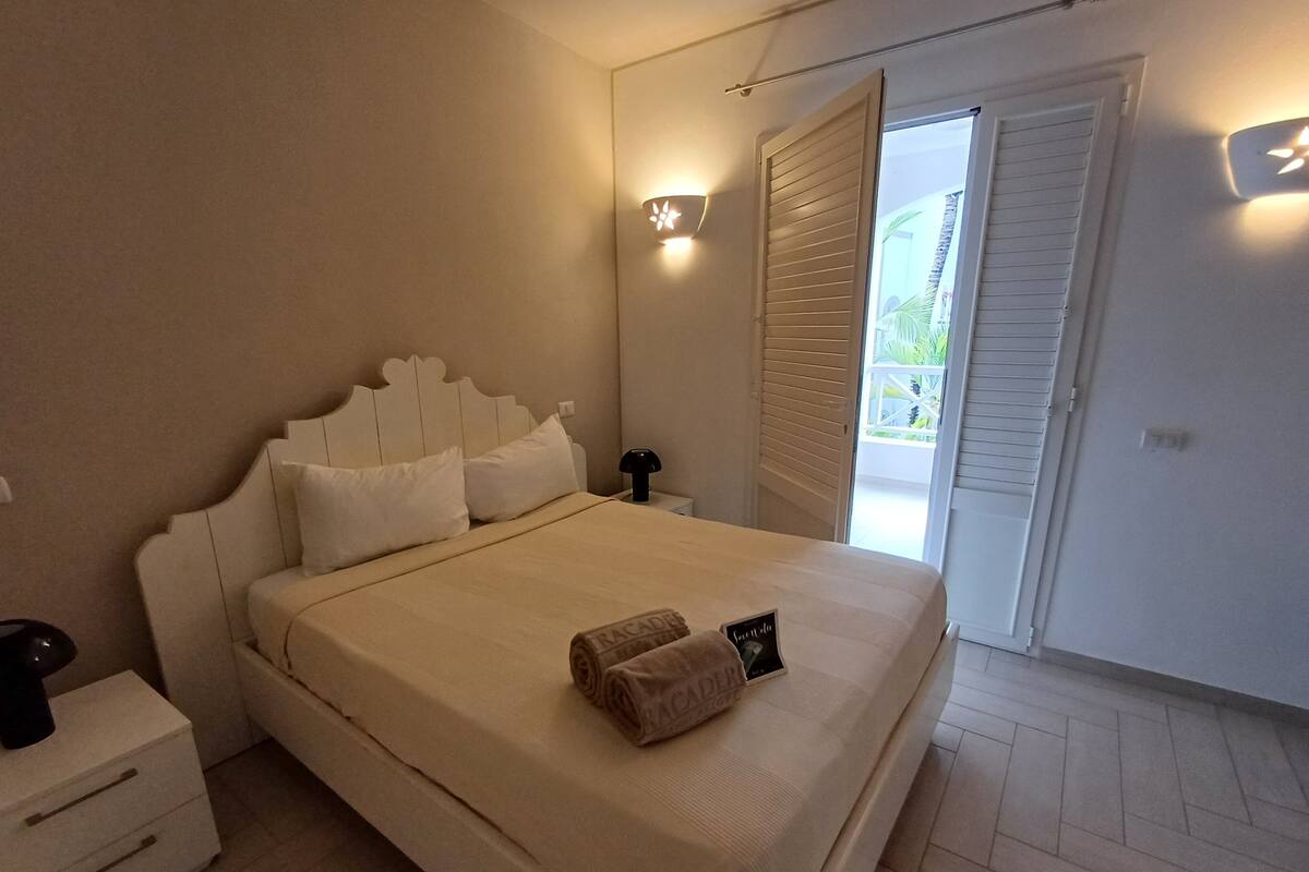 A bedroom with a bed, bedside tables, a nightstand, a wall-mounted light, and a sliding door to an outdoor area.