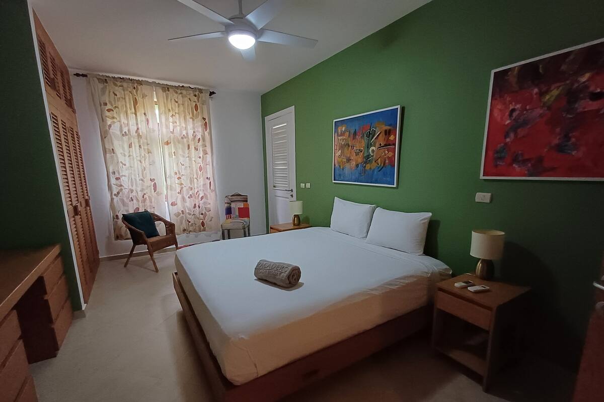 A bedroom with a bed, a chair, a desk, a ceiling fan, and a painting on the wall.