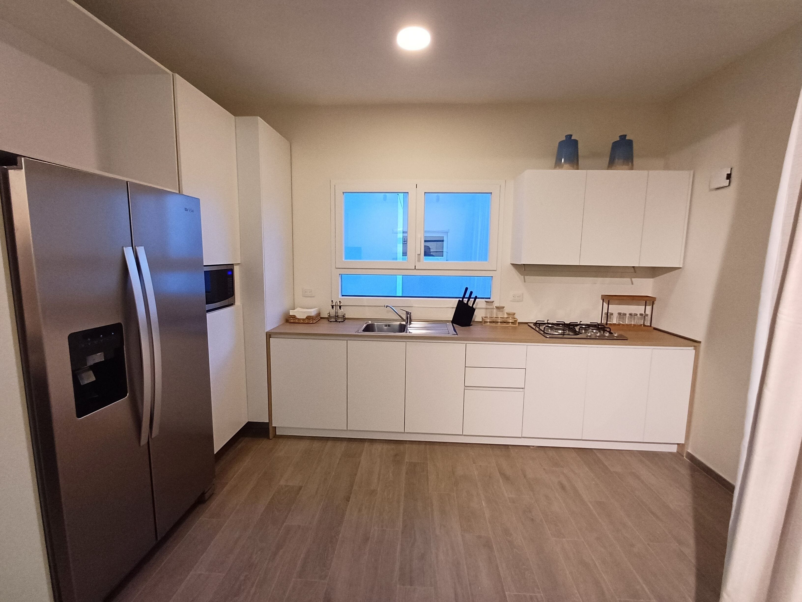 Una cocina moderna con armarios blancos, un refrigerador de acero inoxidable, un lavamanos doble y una ventana sobre el lavamanos.