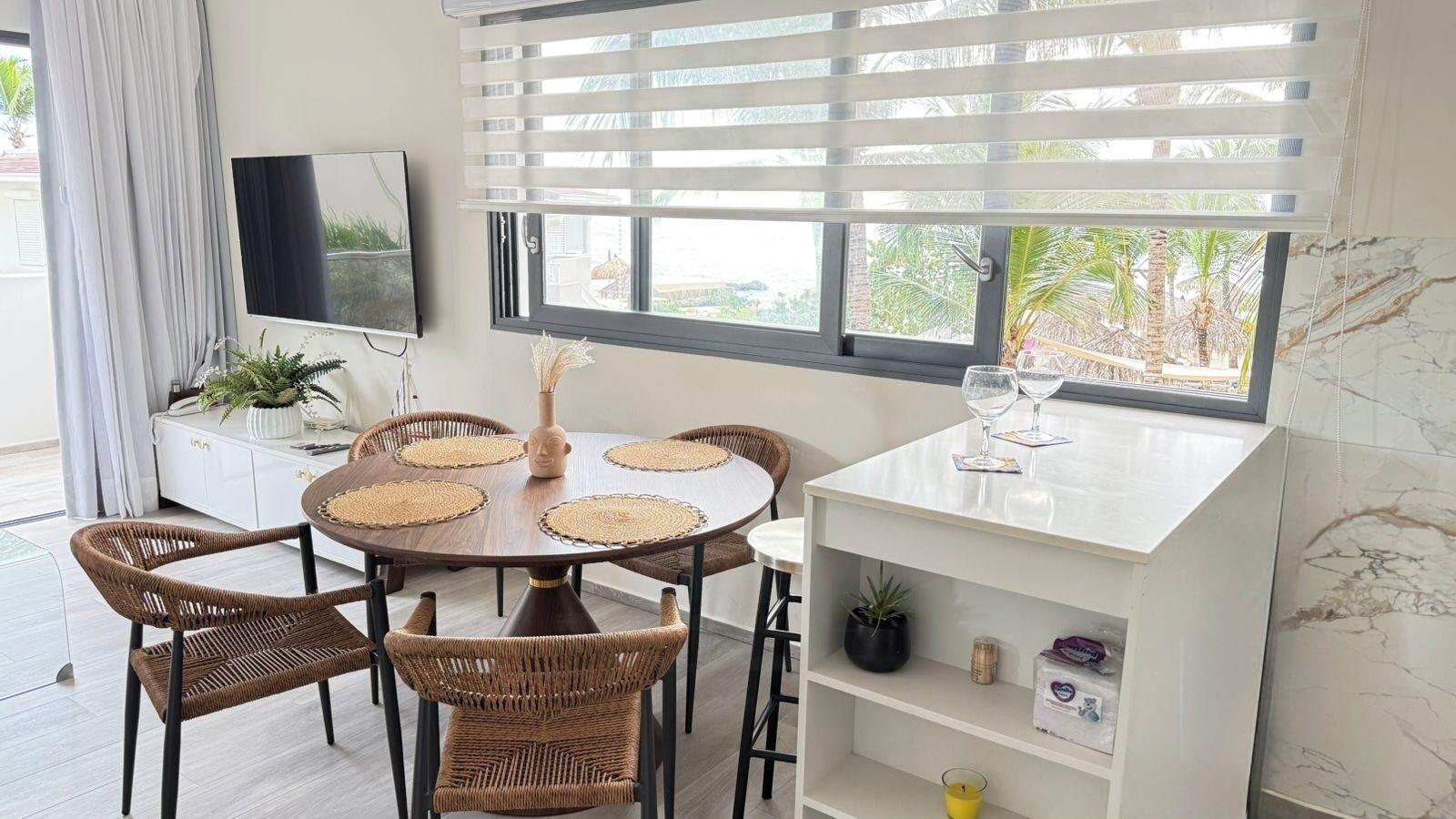 Un coin repas avec une table ronde, des chaises en osier, un meuble blanc, un téléviseur et une fenêtre avec des stores.