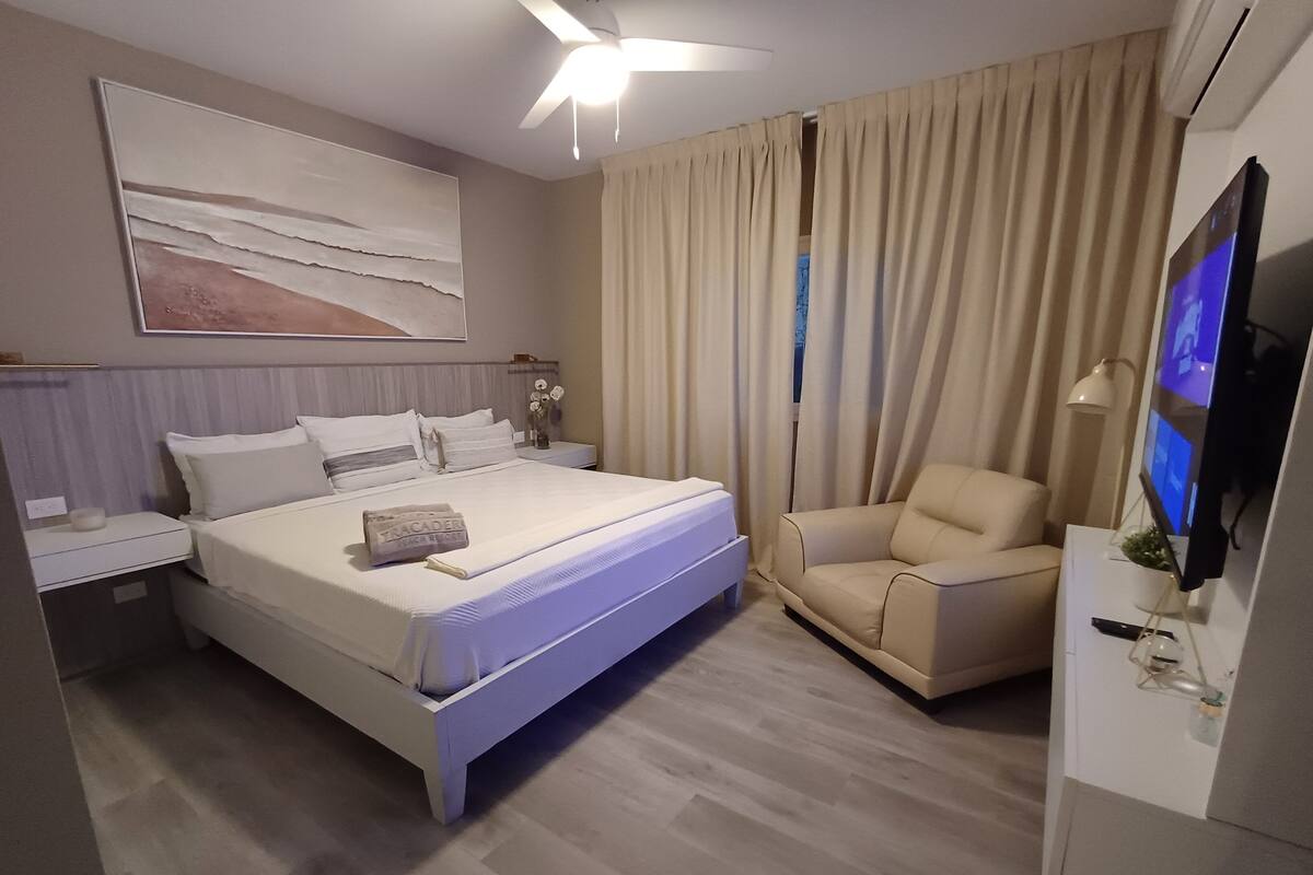 A modern hotel room with a large bed, a chair, a TV, and a ceiling fan.