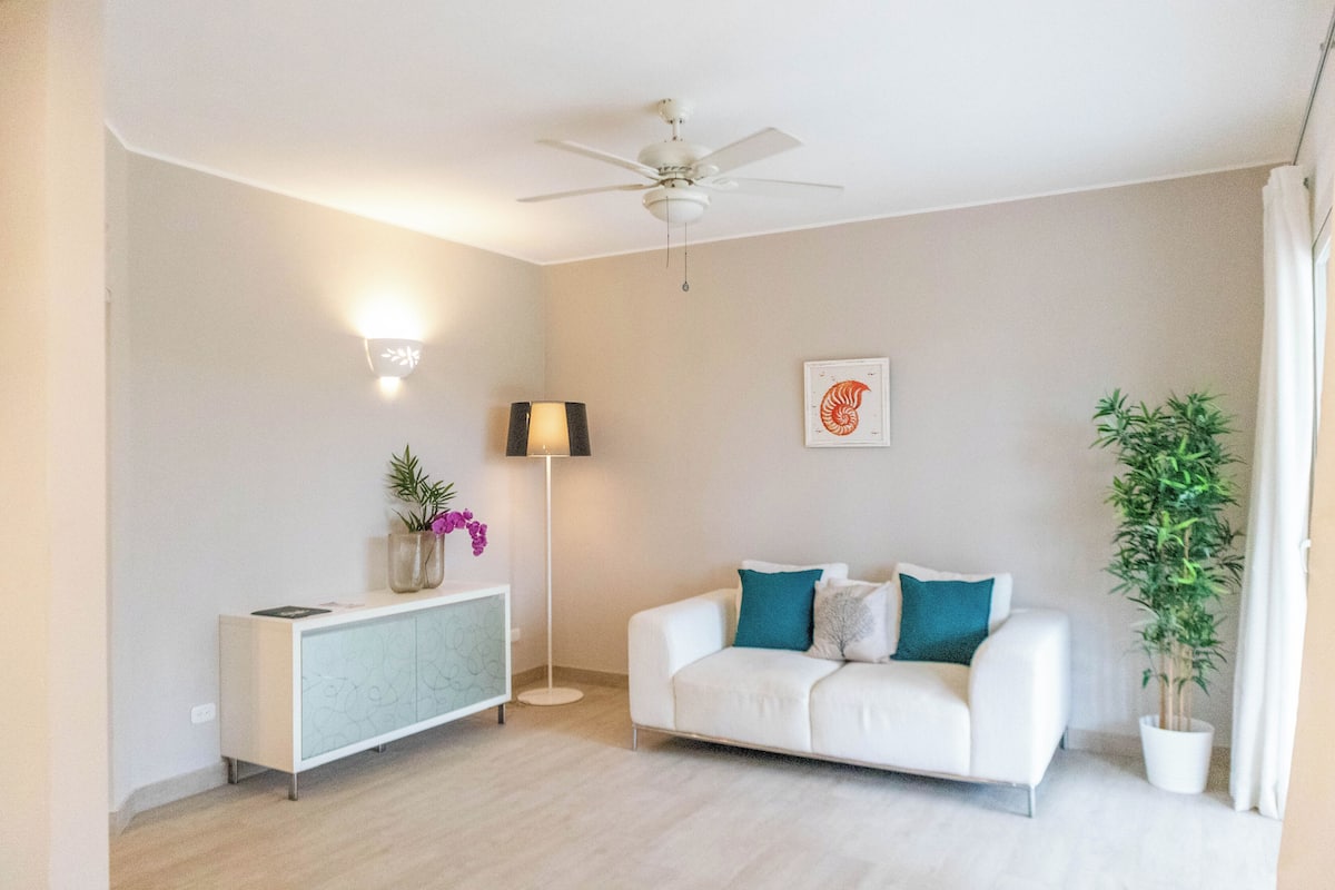 A modern living room with a white sofa, a floor lamp, a ceiling fan, a potted plant, and a decorative wall piece.