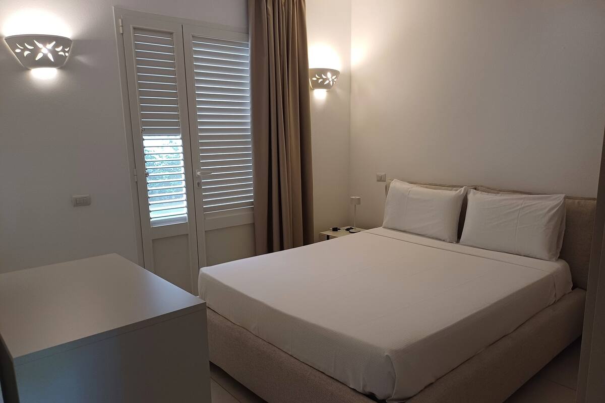 A bedroom with a bed, bedside table, lamp, and window with blinds.