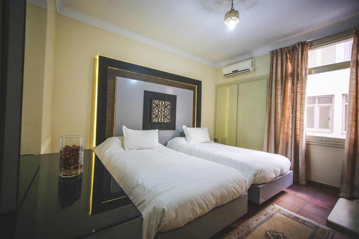 A hotel room with two beds, a glass table with decorative items, a wall-mounted air conditioner, and a window with curtains.