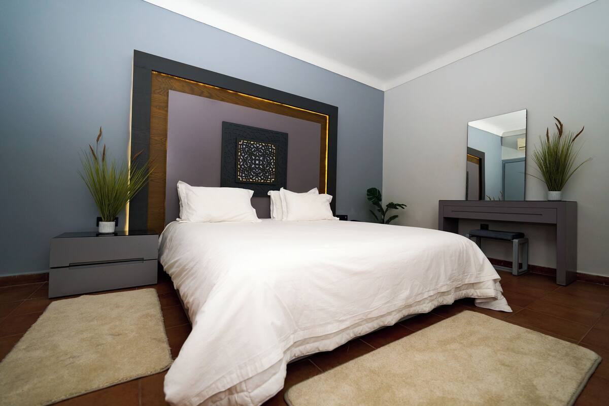 A modern bedroom with a large bed, bedside tables, a dresser, a mirror, and decorative plants.