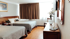 Une chambre d’hôtel avec deux lits, un plancher en bois, une grande fenêtre avec des rideaux, une table de chevet ornée d’un vase de fleurs et un tableau encadré accroché au mur.