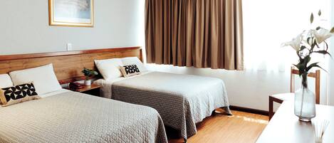 Una habitación de hotel con dos camas, cabecero de madera, mesita de noche y un jarrón con flores, además de una ventana con cortinas.