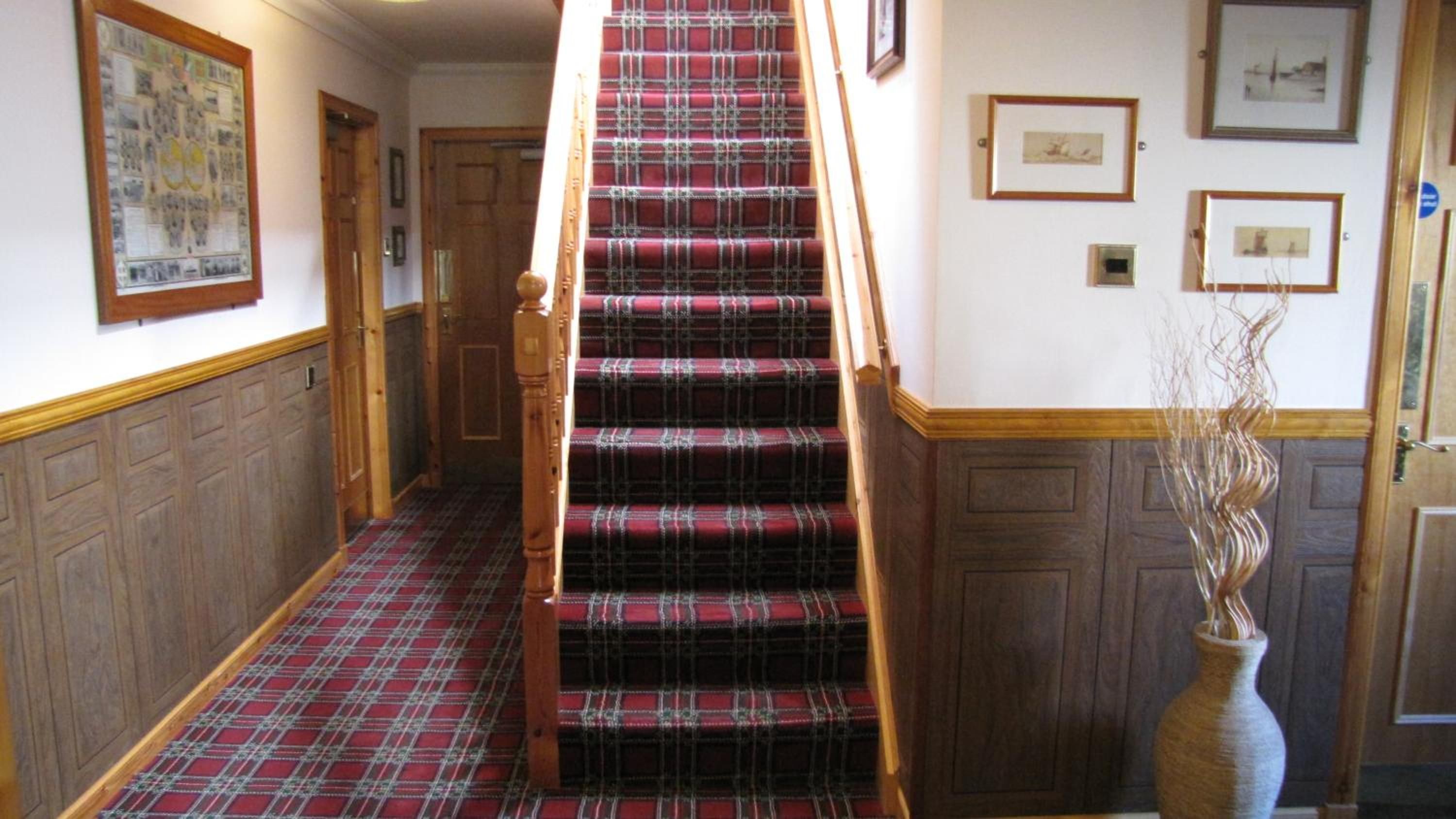 A staircase with a plaid carpet, wooden handrail, and a vase with dried plants.