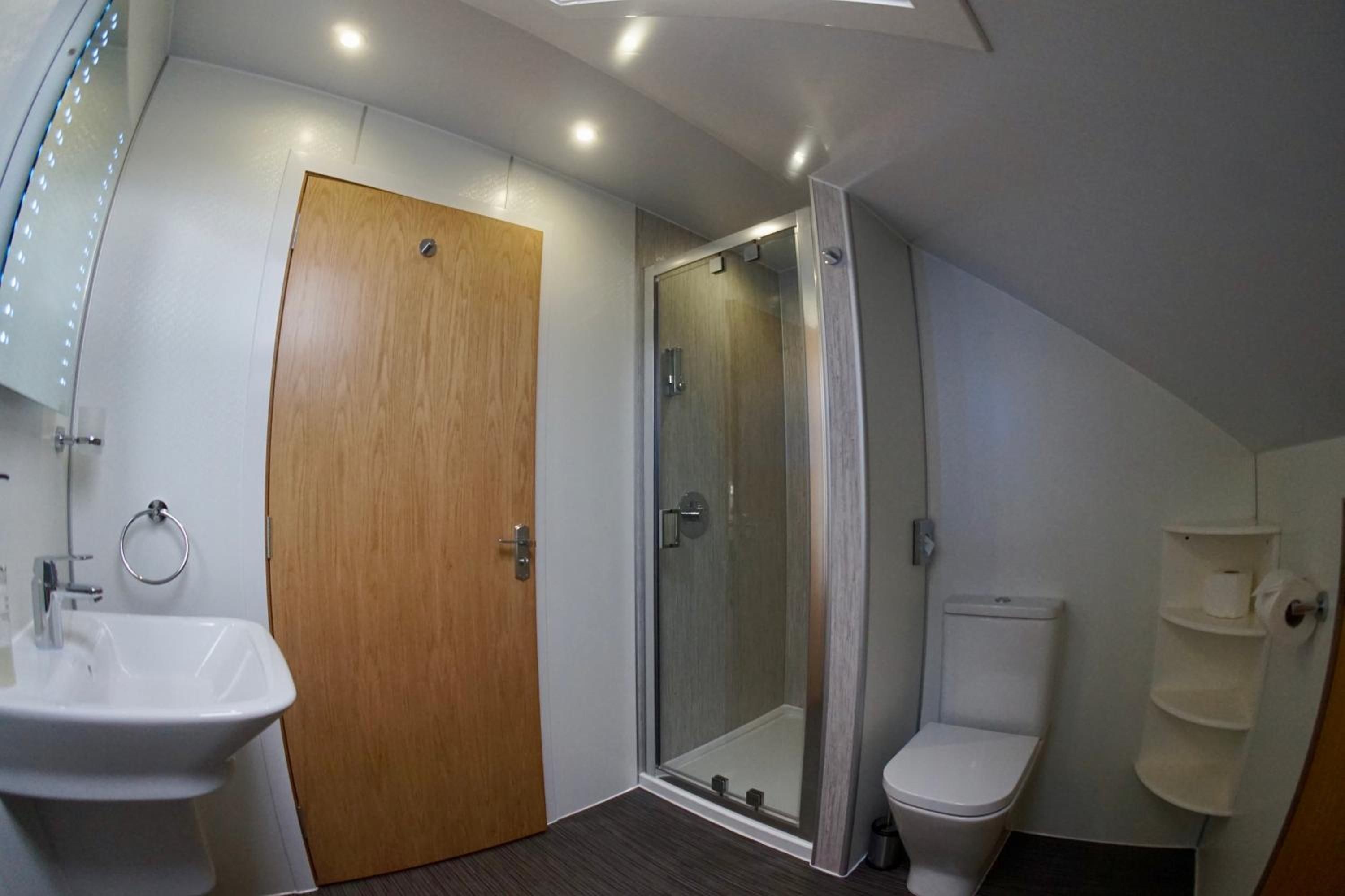 Un baño moderno con puerta de madera, cabina de ducha con mampara de vidrio, lavamanos blanco y un inodoro.
