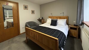 A bedroom with a wooden bed, a chair, a bedside table, a mirror, and a window with curtains. - Karrawa Guest House (Kirkwall)