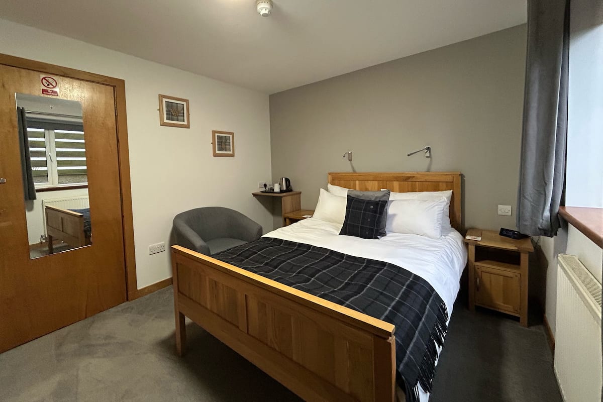 Um quarto com cama de madeira, uma cadeira, mesa de cabeceira, espelho e janela com cortinas.