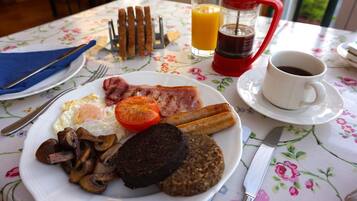 Una mesa de desayuno con un plato de huevos, tocino, champiñones y morcilla, un vaso de zumo de naranja, una cafetera roja y una taza de café.