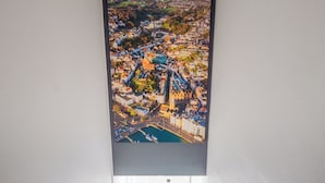Un couloir avec un escalier menant à une porte, une œuvre d’art représentant un paysage urbain encadrée au-dessus de la porte, et une petite plante sur le côté gauche.