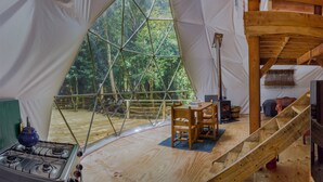 Interior de una cúpula geodésica con suelo de madera, una mesa con sillas, una chimenea y vistas al bosque a través de un ventanal grande.