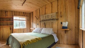 Un dormitorio de cabaña de madera con una cama, un estante con almohadas, un tapiz de pared tejido y una ventana.