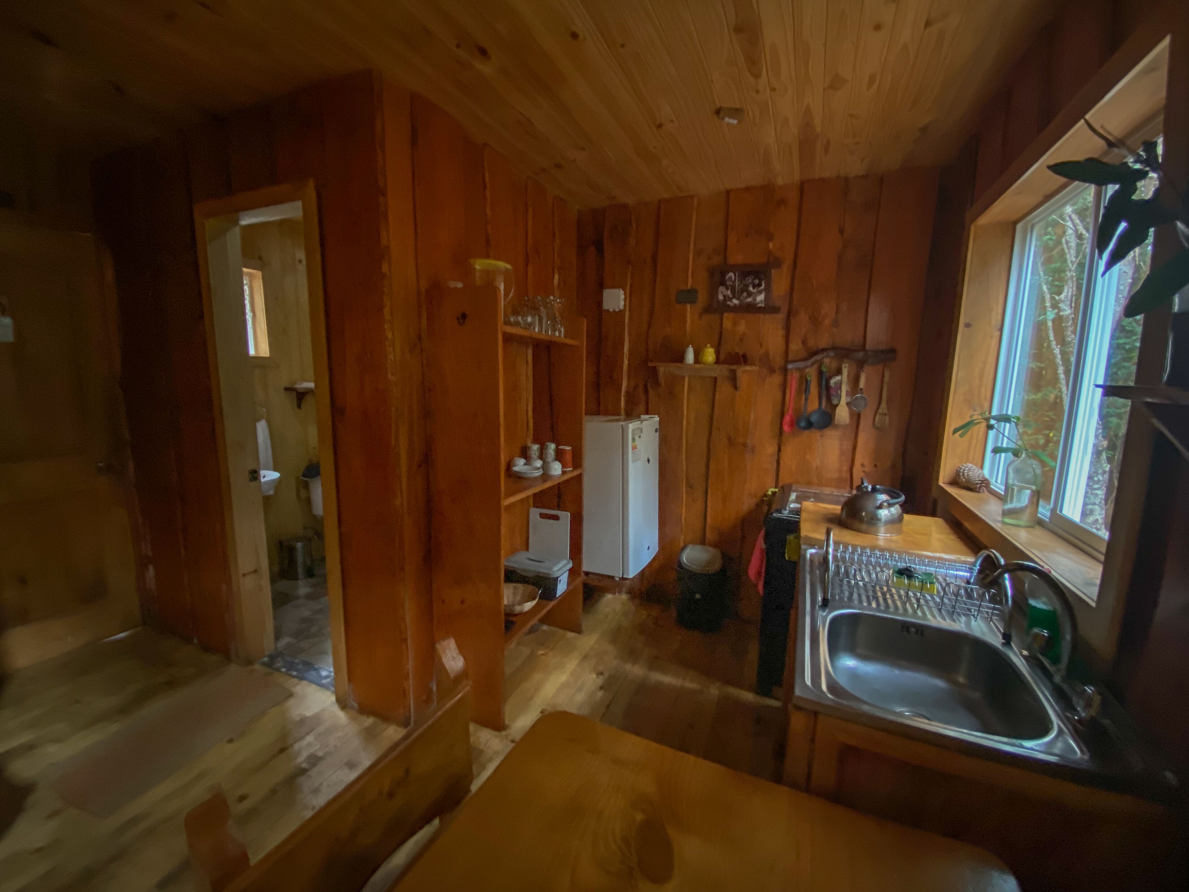 A wooden cabin interior with a kitchen area featuring a sink, a refrigerator, and a dining table.