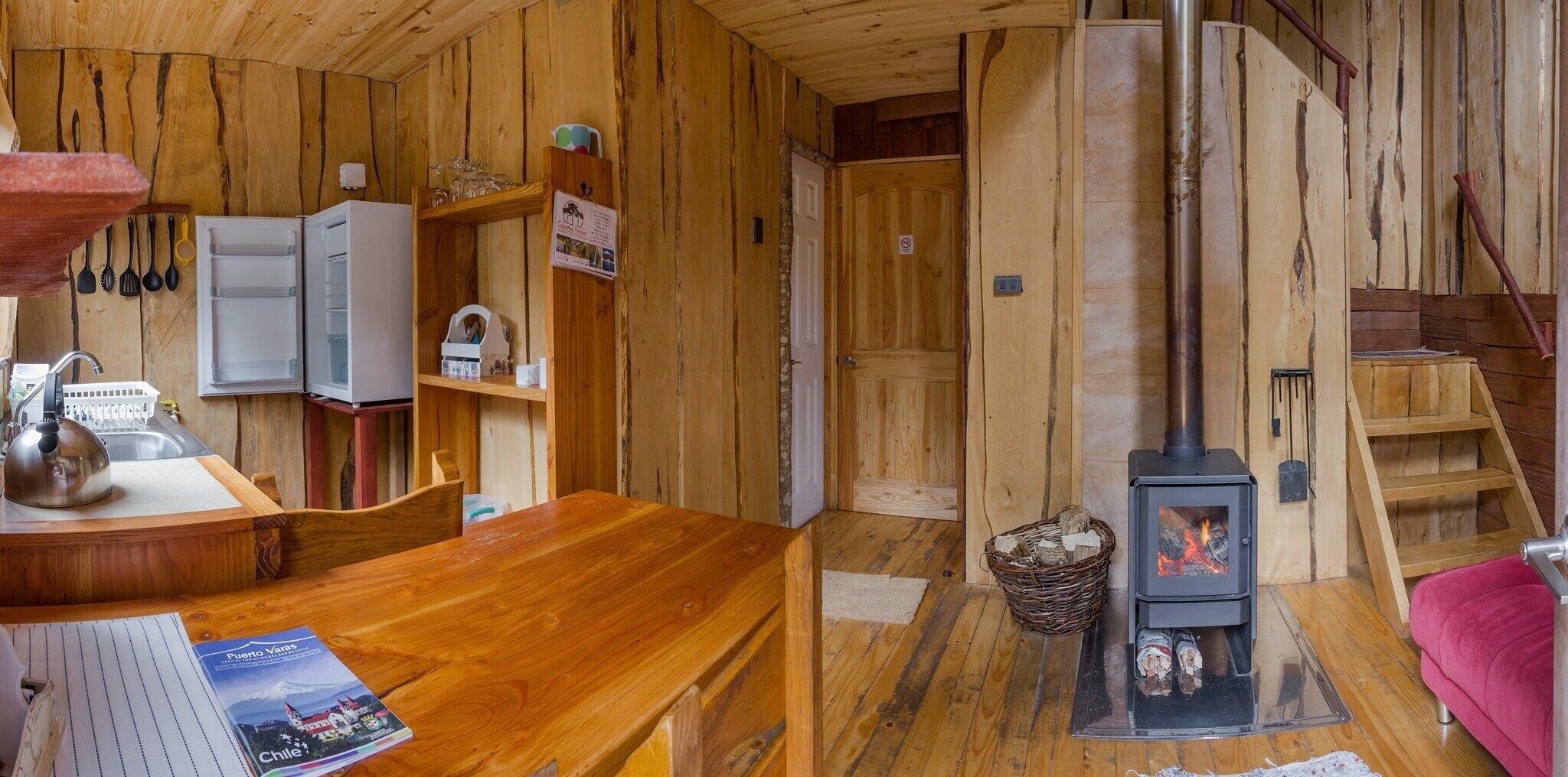 Interior acogedor de una cabaña de madera con comedor, cocina con armarios blancos, escalera de madera y estufa de leña.