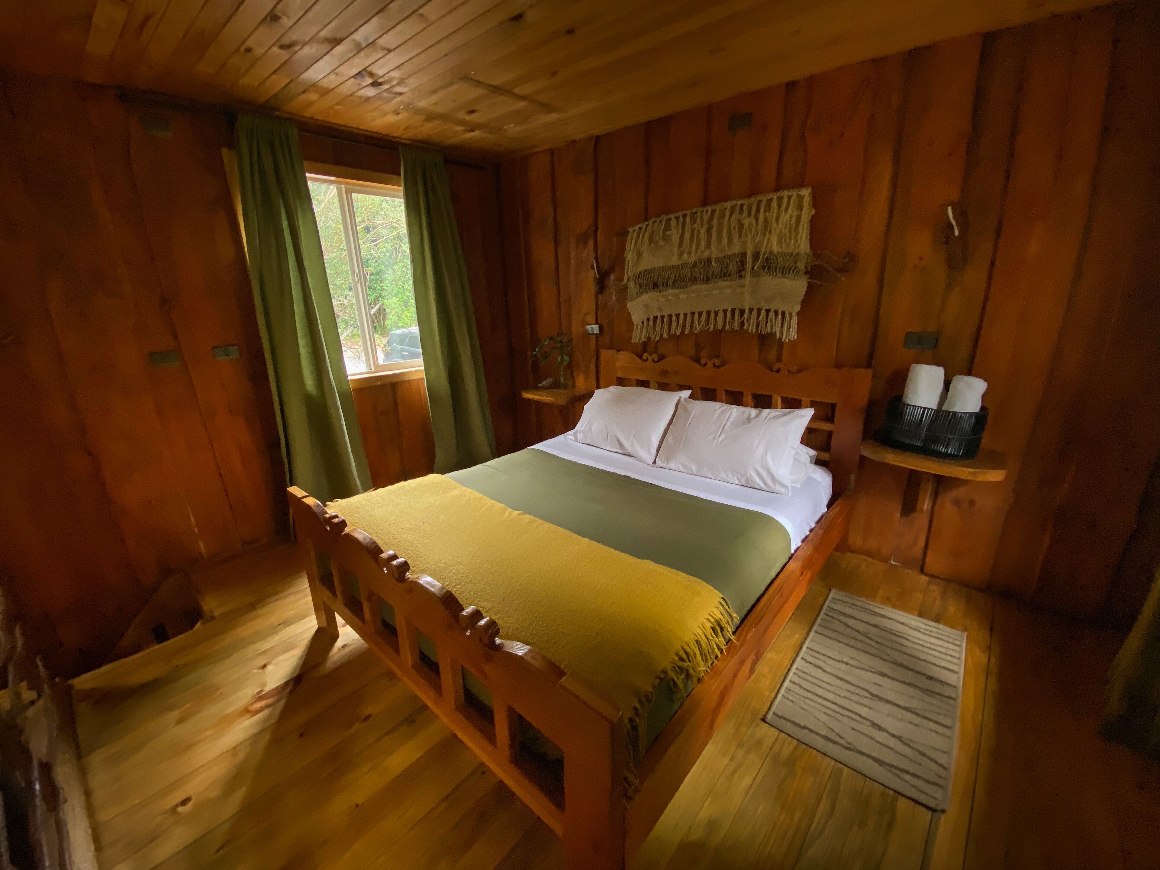 Dormitorio de cabaña de madera con cama, cortinas, cabecera, estante con artículos de aseo y ventana.