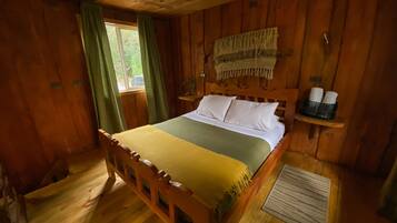 Dormitorio de cabaña de madera con cama, cortinas, cabecera, estante con artículos de aseo y ventana.