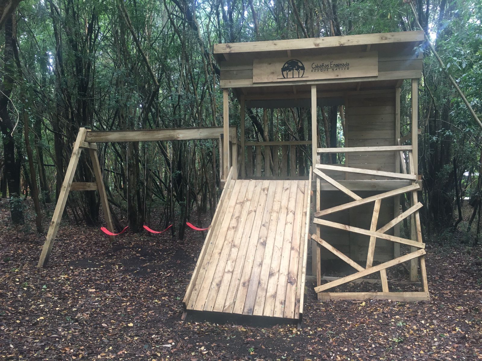 A wooden play structure in a forested area with a sign reading "Cabañas Ensenada SOSQUE NATIVE."