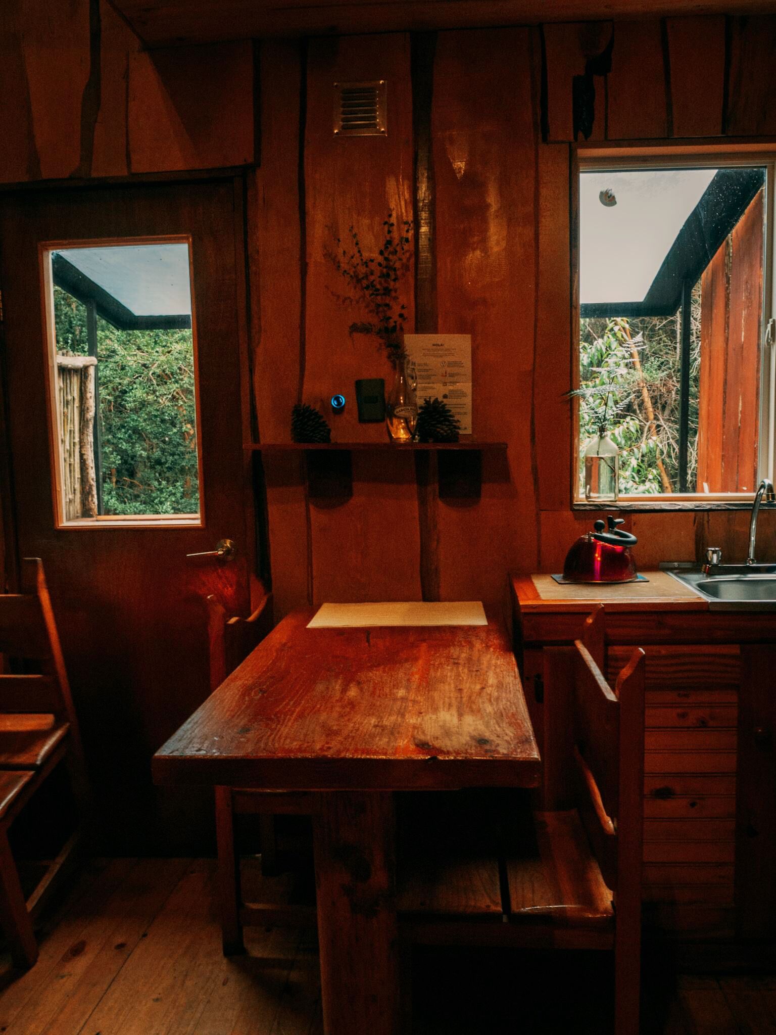 Un interior rústico de madera con una mesa y bancos de madera, una ventana con vistas a la vegetación y un pequeño estante con un jarrón y un menú.