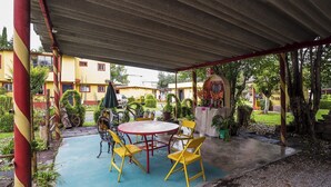 Une terrasse extérieure avec une table ronde et des chaises jaunes, entourée de verdure et abritée par une zone couverte.