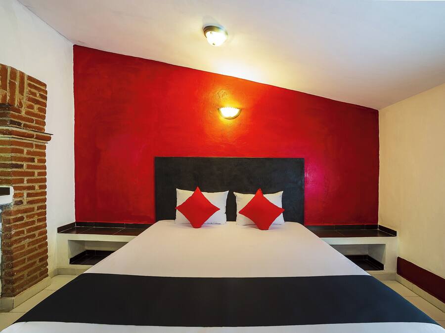 Quarto de hotel com parede de tijolos, parede de destaque vermelha, cabeceira preta, colcha branca e duas almofadas vermelhas.