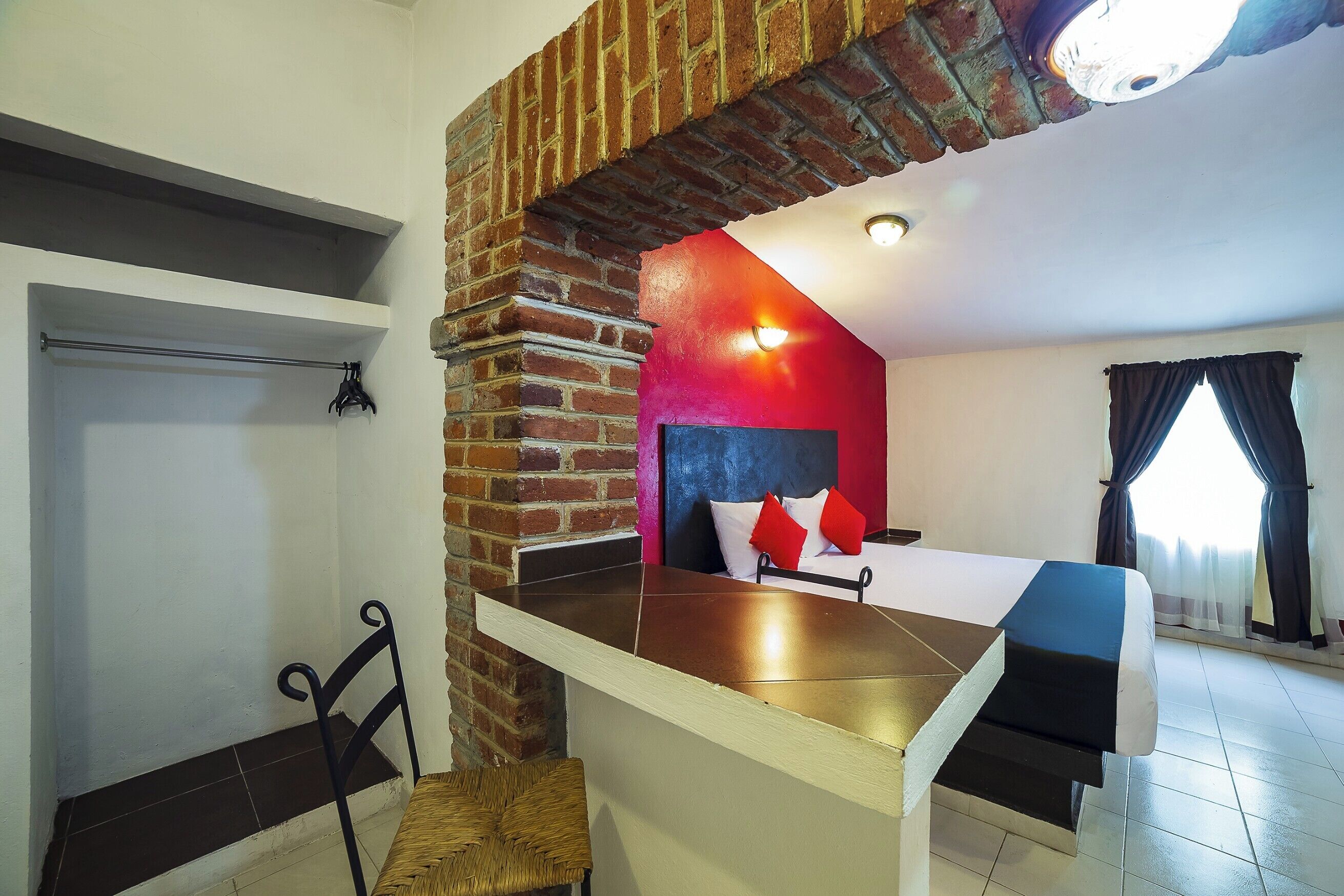 A room with a bed, a small table, a chair, a hanging coat rack, and a brick wall.