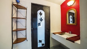 Une salle de bain avec un lavabo blanc, un miroir avec un cadre en bois, une porte noire ornée d’un motif décoratif et une étagère en métal.