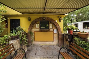 Un espacio al aire libre cubierto con un banco, una jardinera con plantas y una pared amarilla con una ventana y una puerta.