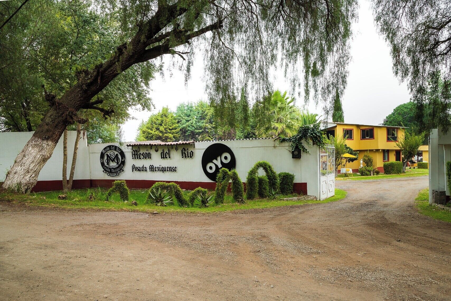 A sign for Meson del Rio Posada Mexiquense with a dirt road leading to a yellow building.