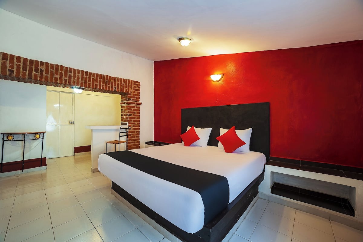 A hotel room with a large bed, a desk, a red accent wall, and a brick fireplace.