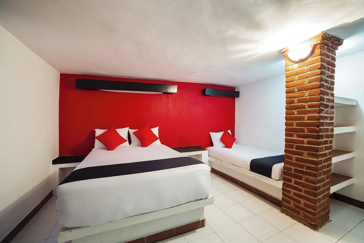 A hotel room with a red accent wall, two beds, a brick pillar, and a white ceiling.
