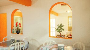 A dining area with a round table and chairs, two arched windows, and a view of a room with a plant and window blinds. - Capital O Hotel Meson Del Rio, Tepotzotlan (Tepotzotlán)